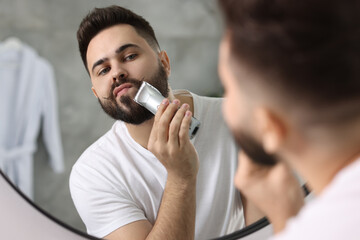 Handsome young man trimming beard near mirror in bathroom