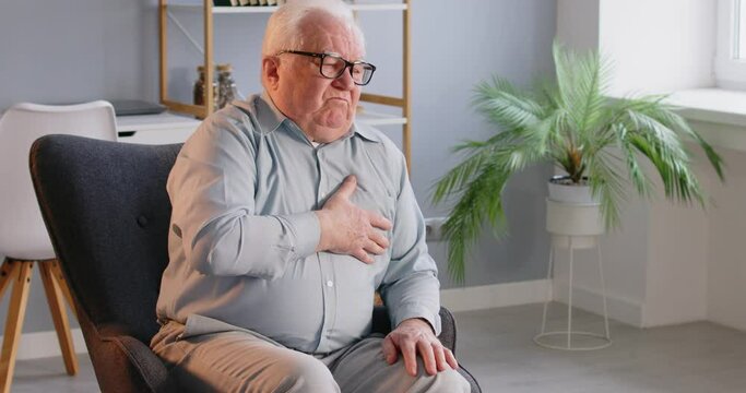 Senior man pressing his chest with hand suffering from heart attack sitting alone on chair at home. Elderly male person having cardiac health problem with myocardial infarction symptoms. 4k video.