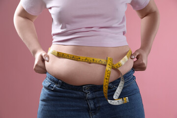 Woman with measuring tape touching belly fat on pink background, closeup. Overweight problem