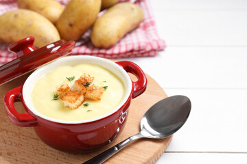 Tasty potato soup with croutons in ceramic pot and spoon on white wooden table