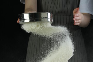 Woman sieving flour at table against black background, closeup