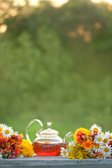glass teapot with herbal tea and colorful flowers on table in garden. Summer nature background....