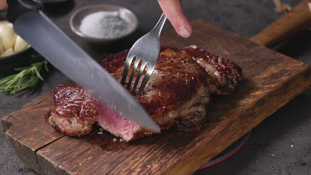Chef slicing freshly grilled juicy beef steak meat with a knife on a wooden cutting board in the kitchen, food close-up