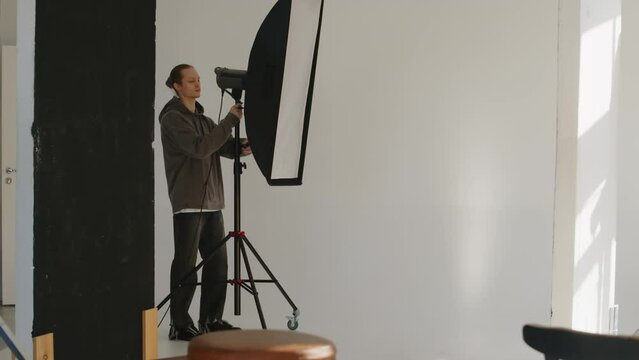 Full shot of young male photographer assistant in relaxed casualwear adjusting softboxes and other lighting equipment while preparing set for studio portrait photoshoot on white background