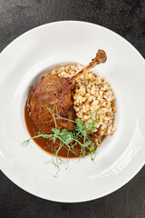 Classic dish french cuisine - roasted duck leg in porto sauce with garnish on black background. Cooked duck leg with pearl barley on white plate on dark concrete table. Duck confit in minimal style.