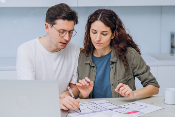 Young couple engages deeply in discussion over home renovation plans, with documents and laptop on their kitchen table. Buying apartment, family mortgage loan. 