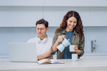 Woman pouring coffee while man works on laptop, both smiling in bright, modern kitchen setting. Happy Couple Sharing Coffee Break While Working on Laptop at Home 