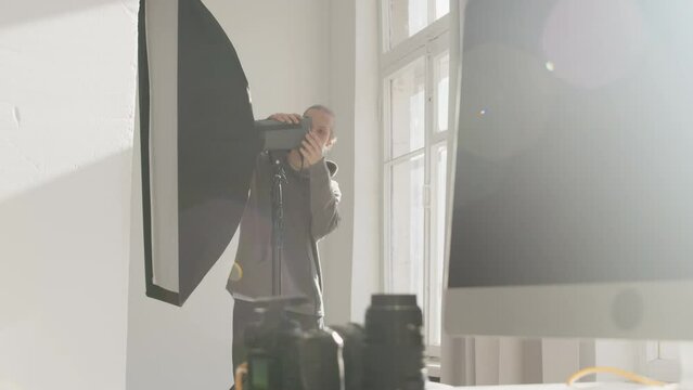 Lens flared shot of young Caucasian male assistant of photographer preparing lighting equipment for having studio portrait photoshoot indoors