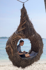 unusual workplace, man working on laptop while sitting in hanging chair nest on the beach