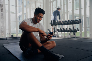 Two multiracial young men in sportswear smiling and using mobile phone next to weights at the gym