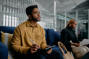 Two multiracial young adults in businesswear sitting on sofa and using digital tablet in office