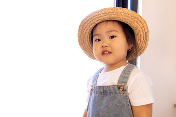 A close-up portrait of a cute little Asian girl. Funny Korean toddler in a straw hat, white T-shirt and denim jumpsuit at home.