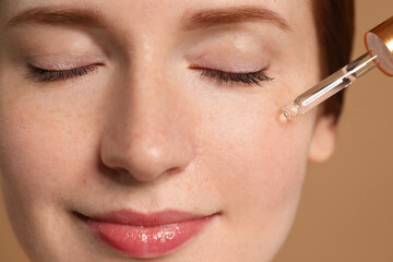 Beautiful woman with freckles applying cosmetic serum onto her face on beige background, closeup