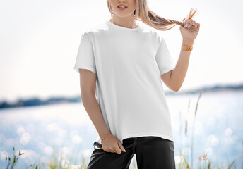 Template of a stylish white T-shirt with a round neck on a blonde girl, posing against the background of a river, front view.