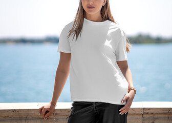 Mockup of a white T-shirt with a round neck on a girl, standing on the embankment, front view.