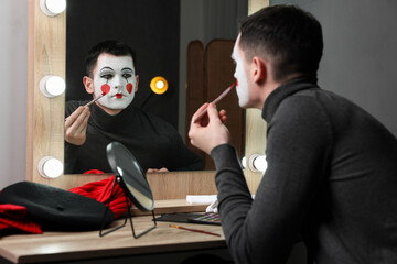 Young man applying mime makeup near mirror in dressing room - Powered by Adobe