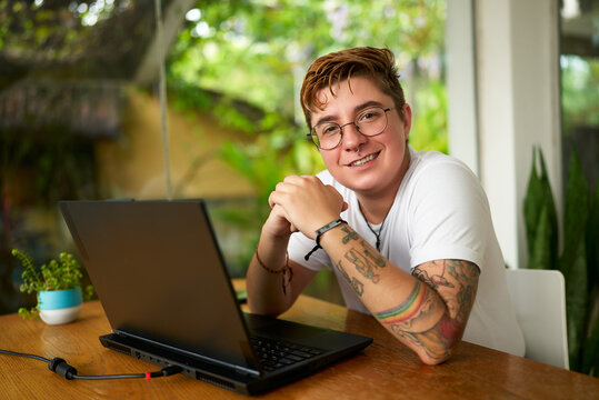 Transgender male person smiles at tropical cafe, laptop on table, earphones nearby. Inclusive work environment, tattoos visible, gender identity embraced, green plants background, youth culture.
