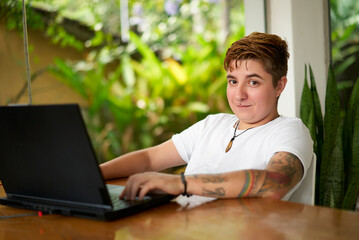 Confident transgender pro works on laptop, casual office setup with green plants. Modern remote job...