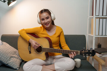 Young woman wears music headphones playing a musical instrument while practicing acoustic guitar and practicing singing sitting on a comfortable sofa in the living room at home.