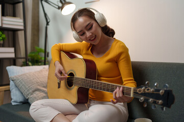 Young woman wears music headphones playing a musical instrument while practicing acoustic guitar and practicing singing sitting on a comfortable sofa in the living room at home.