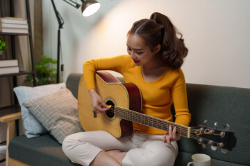 Smiling, happy young woman playing a musical instrument while practicing acoustic guitar and practicing singing sitting on a comfortable sofa in the living room at home.