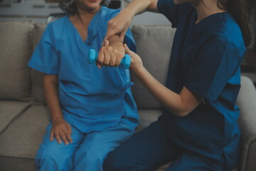 Old woman training with physiotherapist using dumbbells at home. Therapist assisting senior woman with exercises in nursing home. Elderly patient using dumbbells with outstretched arms.
