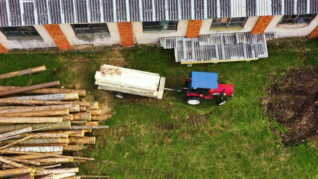 Tractor backs up trailer of milled plank logs on side of sawmill, aerial top down static