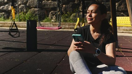Pretty woman checking fitness app on her smartphone, checking heart rate and burnt calories during outdoor workout, sitting on the sportsground nd having a break between sets. Active lifestyle concept