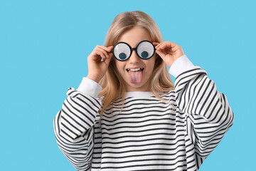 Little girl in funny eyeglasses showing tongue on blue background. April Fools' Day celebration