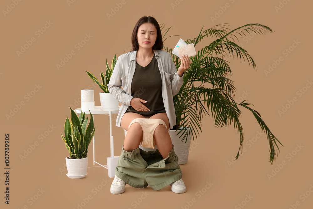 Poster Young Asian woman with menstrual pads sitting on toilet bowl against beige background