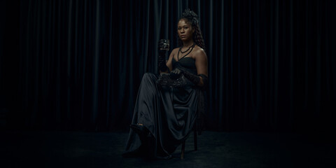 African-American woman, dressed old-fashion dress, looks medieval person, sits confidently on chair holding glass of brandy against dark curtain backdrop. Concept of comparison of eras, vintage. Ad - obrazy, fototapety, plakaty