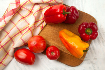 Cutting board with several red and yellow tomatoes and bell pepper on white wooden background with red kitchen towel..