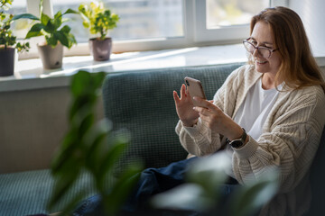 Caucasian attractive mature middle aged woman sitting on the couch at home using smartphone