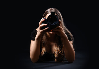 Female photographer with modern camera taking picture of viewer on black background