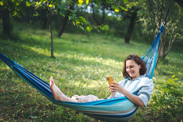 Cheerful young Caucasian woman in a hammock in a summer garden using a smartphone