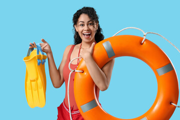 Beautiful young happy African-American female lifeguard with flippers and ring buoy on blue...