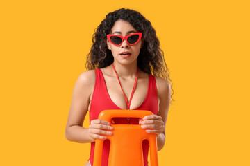 Beautiful young African-American female lifeguard with rescue buoy on yellow background