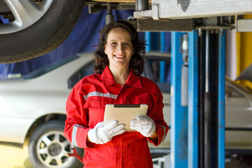 A woman car mechanic stand beneath a lifted car in a garage,  smile while holding tablet computer.