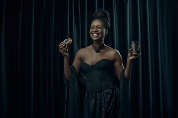Smiling African-American woman, dressed in vintage black dress holds sweet donut and cup of coffee...