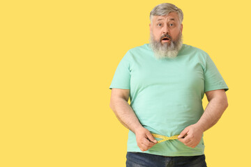 Overweight shocked mature man with measuring tape on yellow background. Weight loss concept