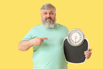 Overweight happy mature man pointing at scales on yellow background. Weight loss concept