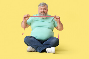 Overweight happy mature man with measuring tape sitting on yellow background. Weight loss concept