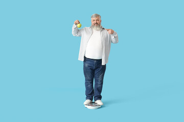 Overweight mature happy man with scales pointing at apple on blue background. Weight loss concept