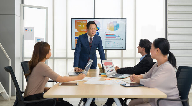 A business meeting in a modern office. A male presenter in suit stand confidently in front of a digital screen displaying colorful chart and graph as if he is delivering a report or presentation. - Powered by Adobe