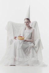 Woman in elaborate white gown and pointed hat, looks as medieval person, sits, holding sweet cake...