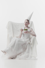 Woman in elaborate white gown and pointed hat, looks as medieval person, sits,eating fresh salad...