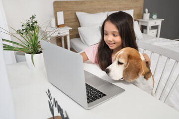 Cute little Asian girl with Beagle dog using laptop at table in bedroom