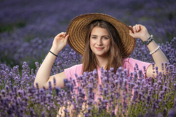 Atractive long haired  tanned girl is posing in a blooming lavender field. holding her straw hat. Horizontally. 