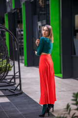 A young woman in bright clothes, a red skirt and a green longsleeve is standing in a business center. High-heeled shoes.