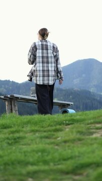 a mother with a baby in an ergo backpack on a hike in the mountains. a mother in sunglasses and a baby in a sling sits on a bench and swings her legs against the background of mountains. vertical vide